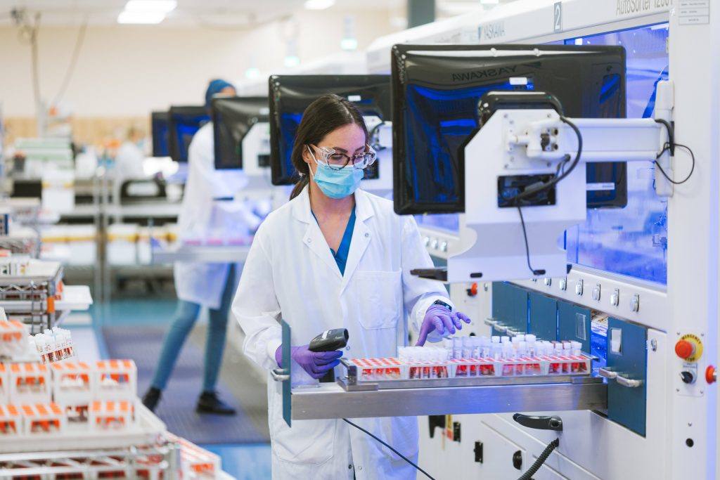 Mayo Clinic Laboratories health care researcher, a white woman wearing glasses and PPE, facemask, gloves while working in the lab with test tubes